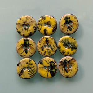 Könnte beinhalten: Ein Set aus neun runden Knöpfen, angeordnet in einem drei-mal-drei Raster. Jeder Knopf hat einen gelben Hintergrund mit detaillierten Illustrationen von Bienen und botanischen Elementen. Jeder Knopf hat zwei mittige Löcher.