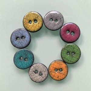 Könnte beinhalten: Ein Set aus acht runden Keramikknöpfen, die in einem Kreis angeordnet sind. Jeder Knopf weist eine ausgeprägte Craquelé-Glasur in Gelb, Grau, Pink, Grün, Orange, Weiß, Blau und Türkis auf. Jeder Knopf hat zwei mittige Löcher zur Befestigung.