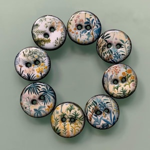 May include: A set of eight circular buttons arranged in a ring, each featuring a glossy white surface decorated with colourful botanical illustrations of various green leaves, blue foliage, and small yellow and orange flowers, with two central holes.