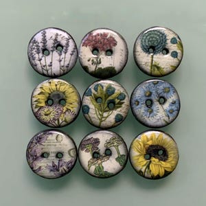May include: A set of nine round, two-hole buttons arranged in a grid. Each button features a unique botanical illustration, including sunflowers, lavender, and various wildflowers, set against a cream-coloured background with faint script text.