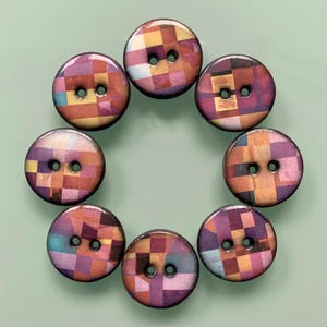 Könnte beinhalten: Eine kreisförmige Anordnung von sieben runden Knöpfen, die jeweils ein Zentrum mit zwei Löchern und ein mehrfarbiges geometrisches Gittermuster in Violett-, Orange-, Gelb- und Hellblautönen mit einer glatten, glänzenden Oberfläche aufweisen.