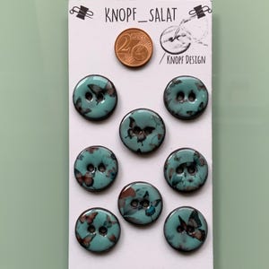 Könnte beinhalten: Acht türkisfarbene Knöpfe mit Schmetterlingsmotiven. Jeder Knopf hat zwei Löcher und eine glänzende Oberfläche. Die Knöpfe sind auf einer weißen Karte mit dem Text "KNOPF_SALAT" und einer 2-Cent-Münze zur Größenreferenz abgebildet.