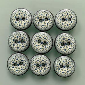 May include: A set of nine round buttons arranged in a three-by-three grid. Each button features a repeating geometric pattern in pale yellow, dark blue, and white, with two central holes for attachment and a dark, solid-coloured outer edge.