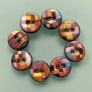 Könnte beinhalten: Ein Set aus acht runden Knöpfen, die in einem Kreis angeordnet sind. Jeder Knopf zeigt ein mehrfarbiges, pixeliges Gittermuster in Orange-, Gelb-, Braun- und Hellblautönen. Jeder Knopf hat zwei mittige Löcher und eine glatte, glänzende Oberfläche.