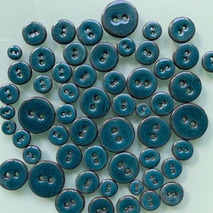 Könnte beinhalten: Eine Sammlung türkisfarbener Knöpfe in verschiedenen Größen. Jeder Knopf hat zwei Löcher und eine glänzende Oberfläche. Die Knöpfe sind auf einer hellgrünen Oberfläche angeordnet und bilden ein ansprechendes Muster.
