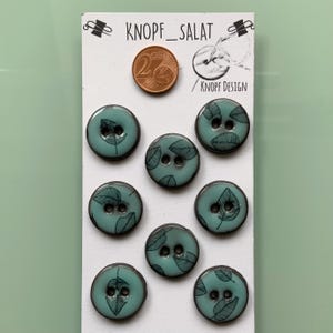 Peut inclure: Ensemble de huit boutons turquoise avec un motif de feuilles, disposés sur une carte blanche. Les boutons ont une finition brillante et deux trous. La carte affiche le texte "KNOPF_SALAT" et une pièce de 2 centimes.