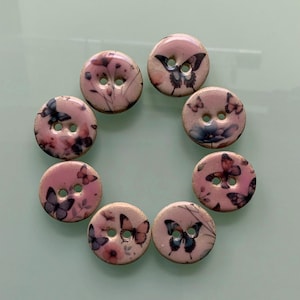 Puede incluir: Ocho botones redondos con diseños de mariposas. Los botones son principalmente rosas, adornados con ilustraciones de mariposas y flores azules, moradas y blancas. Cada botón tiene dos agujeros para coser.