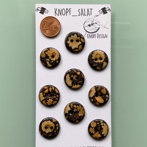 Può includere: Una scheda di nove bottoni decorativi con disegni floreali nei toni del marrone e beige. I bottoni sono rotondi e hanno due o quattro fori. La scheda presenta anche il testo "KNOPF_SALAT" e "KNOPF DESIGN". È visibile anche una moneta da 2 centesimi.