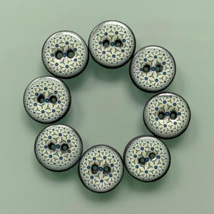 May include: A set of eight circular buttons arranged in a ring. Each button features a cream-coloured surface with a repeating geometric pattern of dark blue dots and light blue star-like shapes. Each button has two central holes for attachment.