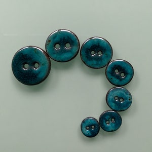 Könnte beinhalten: Ein Satz von sieben tealblauen Keramikknöpfen mit jeweils zwei Löchern. Die Knöpfe sind in einem Halbkreis angeordnet, wobei der größte Knopf oben und der kleinste Knopf unten ist.
