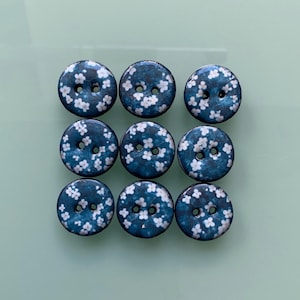 Pode incluir: Nove botões redondos com fundo azul escuro e padrão floral branco. Cada botão tem dois furos para costurar. Os botões estão dispostos em uma grade 3x3.