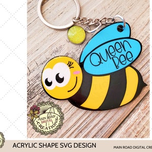 May include: A bee-shaped keyring in yellow and black, with the words "Queen Bee" on a blue wing. It features a silver ring and a small, glittery charm. The design is an acrylic shape SVG.