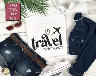 Pode incluir: Uma camiseta branca com um design gráfico preto que diz "travel is my therapy" com um avião e um coração. A camiseta está colocada sobre uma superfície de madeira branca com um par de shorts jeans azuis, um cinto marrom, óculos de sol, uma jaqueta jeans e fones de ouvido brancos.