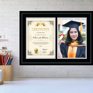 May include: A black framed display featuring a graduation photograph and a certificate. The certificate has gold detailing and text. The photograph shows a graduate in a black cap and gown holding a rolled diploma. The frame is on a white brick wall.