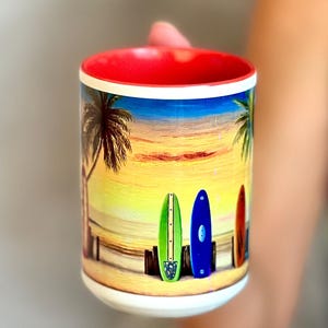 May include: A white ceramic mug with a red rim and a colorful image of palm trees and surfboards against a sunset background.