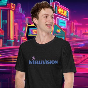 May include: A dark heather grey t-shirt with a blue and purple logo that says "Intellivision" and a pixelated running figure. The background is a blurry image of a neon cityscape.