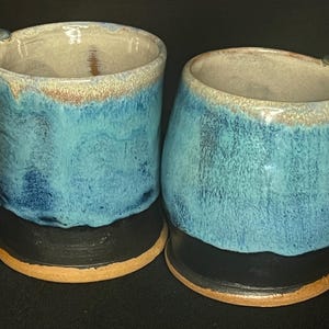 以下が含まれることがあります： 青と黒の釉薬をかけた手作りセラミックマグカップ2個。マグカップはユニークなハンドルが付いており、コーヒーや紅茶に最適です。