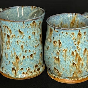 以下が含まれることがあります： 青と茶色の釉薬をかけた、手作りでできたセラミック製のマグカップ2つ。マグカップは独特の斑点模様で、片方は二重の取っ手があります。