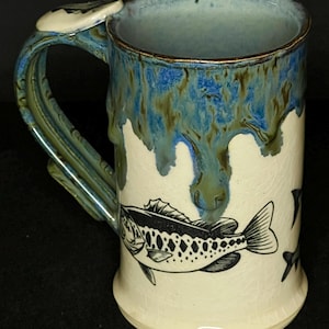 May include: Handmade ceramic mug with a unique design. The mug features a black and white fish illustration on a cream-colored background. The handle and rim have blue and green glaze, with a drip effect. A functional and decorative piece.
