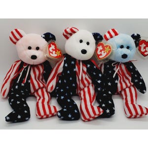 May include: Three plush teddy bears with patriotic designs. The bears have white, pink, and light blue heads, with red and white striped ears. Their bodies are black with white stars, and red and white striped legs.