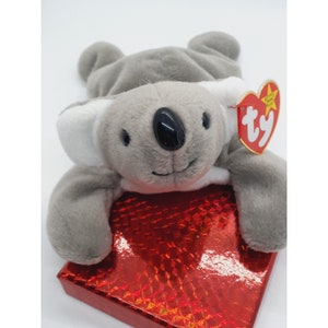 May include: Gray and white plush koala bear with a red Ty tag that says "Ty" and "Beanie Babies" on a red sparkly surface.