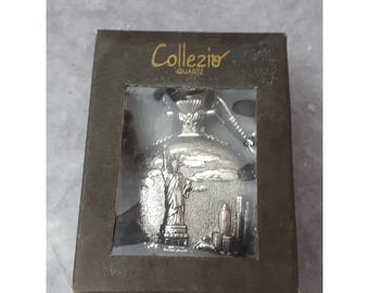 May include: A silver pocket watch with a chain, featuring a detailed engraving of the Statue of Liberty and the New York City skyline. The watch is in a black box with the word "Collezio Quartz" printed on the top.