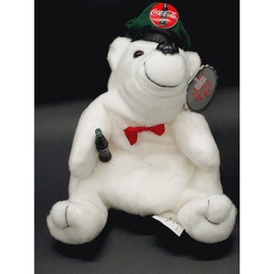 May include: White plush polar bear wearing a green and black hat with a Coca-Cola logo. The bear is holding a miniature Coca-Cola bottle and wearing a red bow tie. The bear has a tag that says "Coca-Cola Polar Bear Plush"
