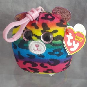 May include: A colorful plush toy in the shape of a bear with a rainbow pattern and leopard print spots. It has large, glittery eyes, a pink nose, and a pink clip. A heart-shaped tag with the words "Squishy Beanies" is attached.