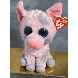 May include: A plush pig toy with soft pink and light purple fur, large glittery blue eyes, and a pink snout. The stuffed animal features silver glittery hooves and a heart-shaped red Ty Beanie Boos tag attached to the ear, displaying the brand logo.