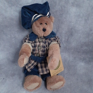 May include: A plush teddy bear wearing a blue corduroy hat with white stripes and a matching outfit. The bear's outfit includes a plaid shirt, blue pants, and a blue belt. The bear has a tag attached to its arm.