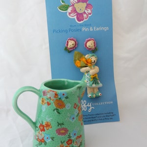 May include: A green ceramic pitcher with a floral pattern. The pitcher has a handle and a spout. The pitcher is sitting on a white surface. The image also shows a card with a bear holding flowers and the text "Mum's the Word Picking Posies Pin & Earrings By COLLECTION".