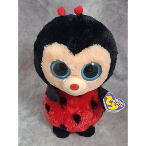 May include: A plush ladybug toy with a round black head, large blue eyes, and a red body featuring black spots. The toy has two small red antennae on top and a small white muzzle with a red nose. A heart-shaped Ty Beanie Boos tag is attached.