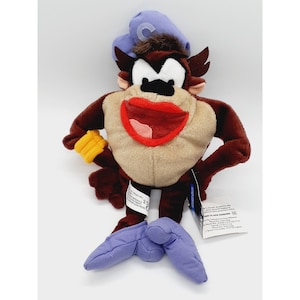 May include: A plush toy of the Tasmanian Devil, a cartoon character. The toy is brown with a tan belly, large red mouth, and a blue hat. It has yellow gloves and purple feet. A tag is attached.