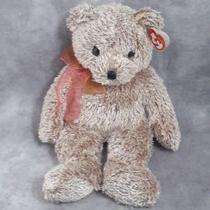 May include: A plush teddy bear with light brown fur, black eyes, and a black nose. It has a red ribbon tied around its neck and a Ty tag attached to its ear. The bear is sitting upright.