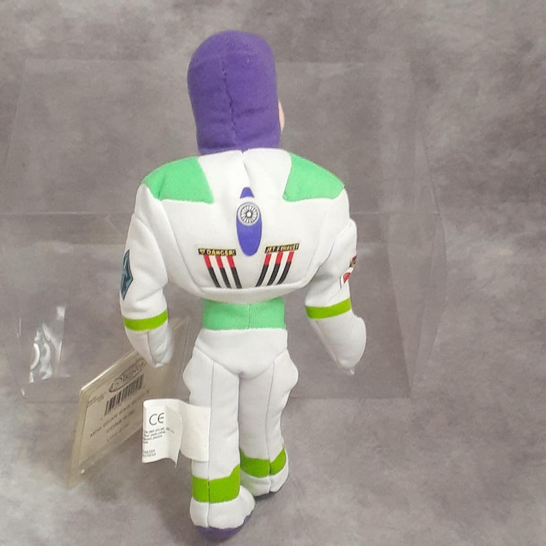 May include: A plush Buzz Lightyear toy, viewed from the back, featuring a purple head, white spacesuit with green and black accents. The toy has a tag attached. The toy is approximately 8 inches tall.