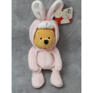 May include: A plush Winnie the Pooh bear wearing a pink bunny costume with long ears. The bear is sitting with its legs crossed. The tag reads "The Disney Store".