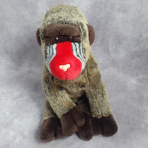 May include: A plush toy baboon with a detailed face. The baboon has a red nose, a white snout, and a face with blue and black stripes. The baboon's fur is brown and tan, with dark brown hands and feet. The toy is sitting upright.