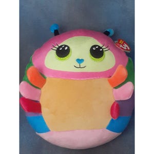 May include: A colorful plush toy shaped like a bug with large, expressive eyes. The toy features a pink head, green face, and a yellow body. It has orange and pink appendages, and a rainbow-colored bottom. A small tag is attached.