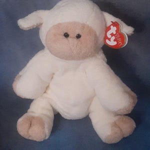 May include: A plush white lamb stuffed animal with a light brown face, ears, and feet. The lamb has black button eyes and a red heart-shaped tag that reads "Pluffies". The toy is soft and cuddly.