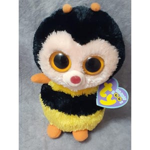 May include: A black and yellow bee plush toy with large, bright eyes. The toy has a pink nose and a Ty Beanie Boo tag attached to its side.