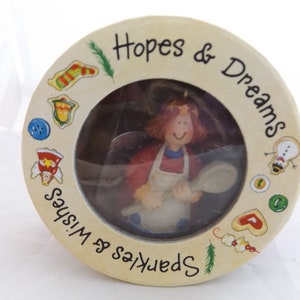 May include: A round, cream-colored ornament with a clear window showing a small figurine of a woman holding a wooden spoon. The ornament is decorated with a variety of festive images, including a snowman, a stocking, and a gingerbread cookie. The text "Sparkles & Wishes" is written on the left side of the ornament, and the text "Hopes & Dreams" is written on the right side.