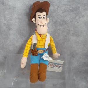 May include: A plush toy of the character Woody from the Disney movie Toy Story. Woody is wearing a brown cowboy hat, yellow plaid shirt, blue jeans, and brown boots. He has a white vest with a cowhide pattern and a brown holster. The Disney Store tag is attached.