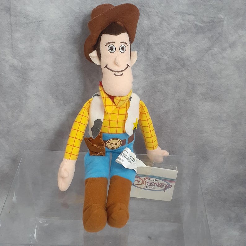 May include: A plush toy of the character Woody from the Disney movie Toy Story. Woody is wearing a brown cowboy hat, yellow plaid shirt, blue jeans, and brown boots. He has a white vest with a cowhide pattern and a brown holster. The Disney Store tag is attached.