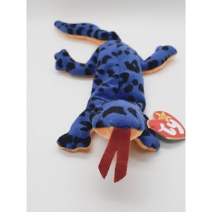 May include: Blue and black spotted plush toy lizard with a red tongue. The toy has a Ty Beanie Baby tag attached to its arm.