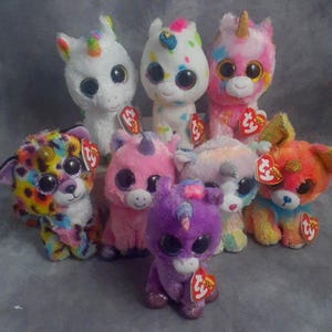 May include: A collection of plush unicorn and animal toys in various colors. The toys have large, glittery eyes and colorful accents, including rainbow horns and spots. Each toy has a red tag attached.