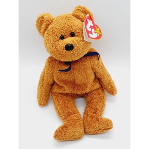 May include: A brown plush teddy bear with a blue ribbon around its neck. The bear has a Ty tag attached to its ear with the text "Ty" and a star.