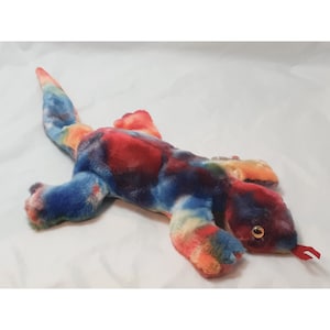 May include: A colorful tie-dye plush lizard toy with a red tongue. The lizard is lying on its belly with its head turned to the side.