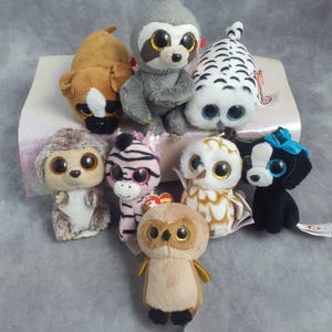 May include: A collection of plush animal toys, including a brown dog, a gray sloth, a white owl with black markings, a hedgehog, a pink zebra, a white and brown owl, a black dog with a blue bow, and a brown owl. Each toy has large, glittery eyes.