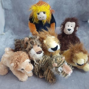 May include: A collection of plush animal toys, including a monkey, lions, a tiger, and a leopard. The monkey has yellow hair and a blue shirt. The lions and tiger have brown and tan fur. The leopard has brown spots.