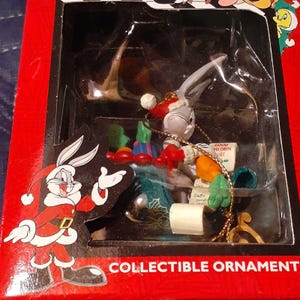 May include: A Bugs Bunny Christmas ornament featuring Bugs dressed as Santa Claus, pulling a sleigh with a 'Good Children List' and a 'Dear Santa' letter. The ornament is packaged in a red box with the words 'Collectible Ornament' printed on the bottom.
